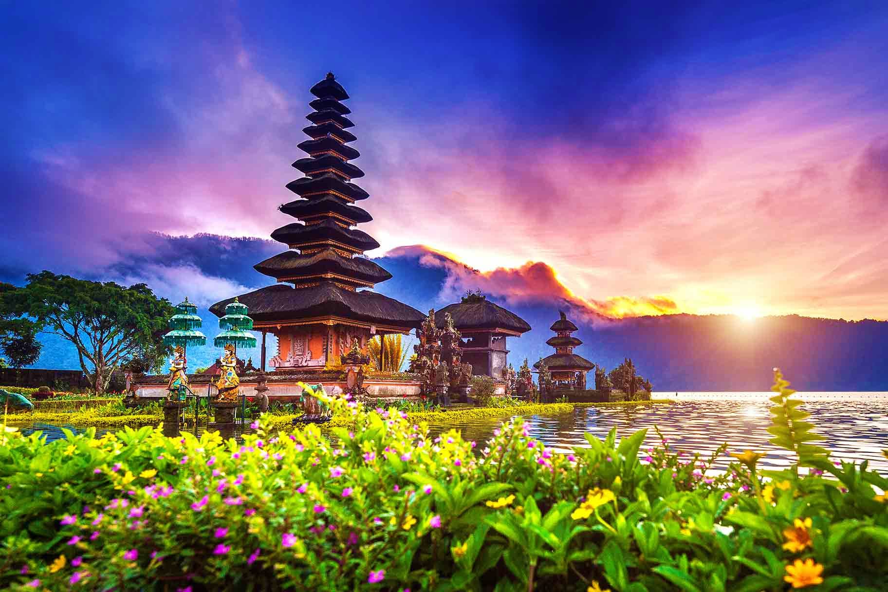 Indonesia Visa Historic Balinese temple with tiered roofs reflected in lake at sunrise, surrounded by colorful flowers and mountains.